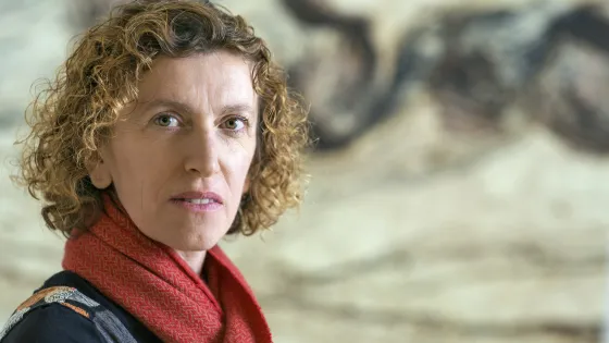 A woman with curly hair and a red scarf stands in front of a blurred artistic background.