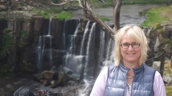 A smiling woman with blonde hair wearing glasses and a blue vest stands in front of a scenic waterfall.