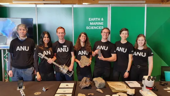Group of seven individuals wearing black ANU T-shirts standing behind a table with geological samples at an Earth and Marine Sciences event.