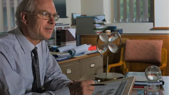 A man sits at his desk in a cluttered office, smiling as he looks at something to the side, with scientific gadgets and papers around him.