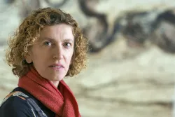 A woman with curly hair and a red scarf stands in front of a blurred artistic background.