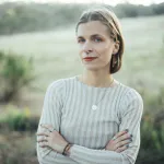 A researcher standing outdoors with arms crossed, wearing a striped shirt and minimal jewellery, looking at the camera against a blurred natural background